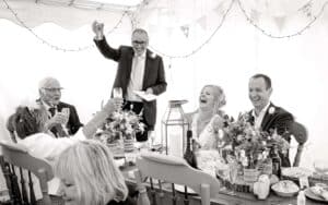 father of the groom delivering a wedding speech and toast. Guests hold glasses in the air.