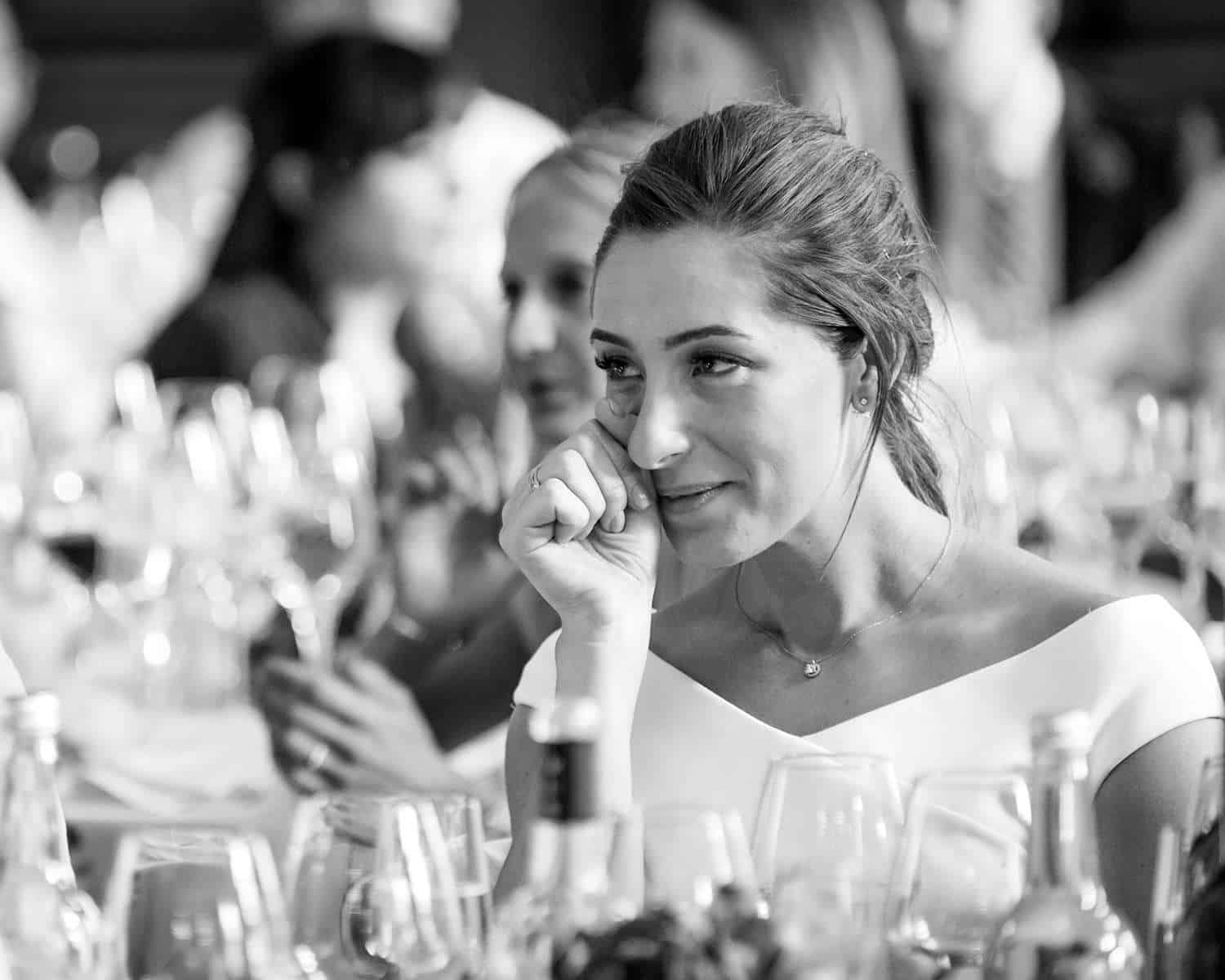 Bride is sitting listening to the groom speech and wiping away a tear.
