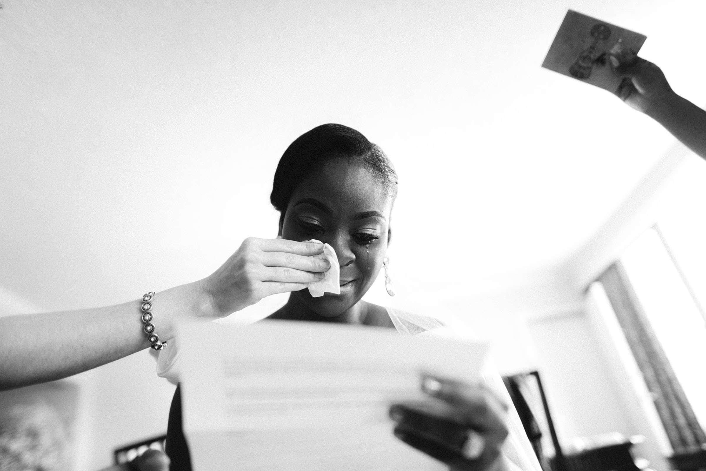 Black bride is reading a letter on her wedding day. She is crying and we can see someone's arm holding a tissue and wiping away her tears.