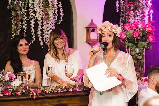 Lesbian bride delivering a wedding speech. She is holding a microphone and her bride and friend are watching and smiling.