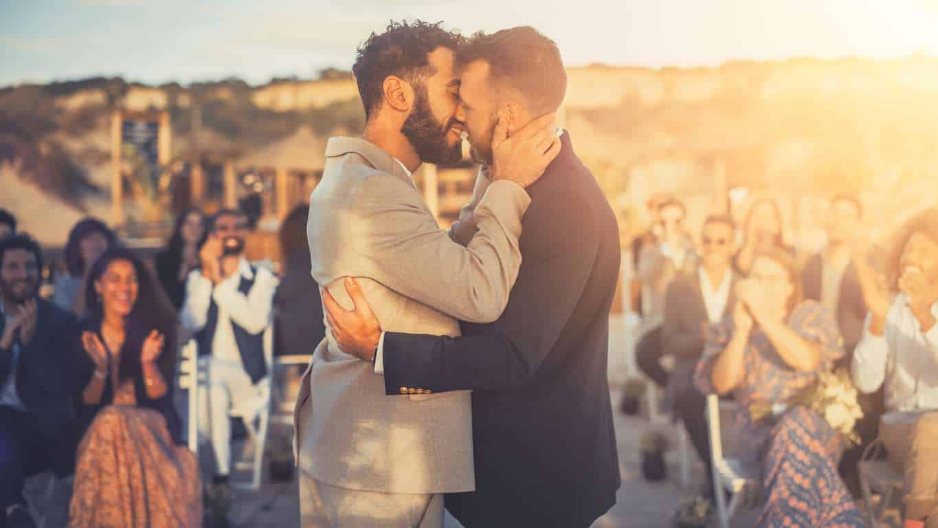 Gay grooms hugging and kissing each other whilst guests clap in celebration.