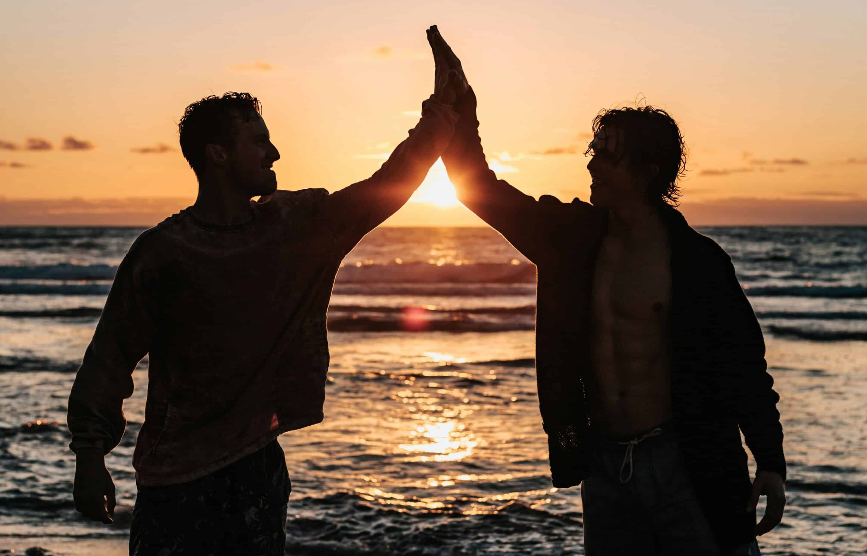 Gay grooms doing a high five by the sea with the sun setting.